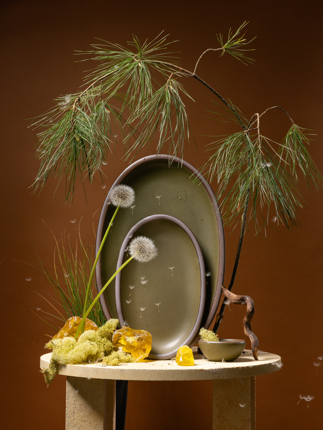 Decorative setting with ceramic oval platters in olive green, amber stones, and a plant on a brown background