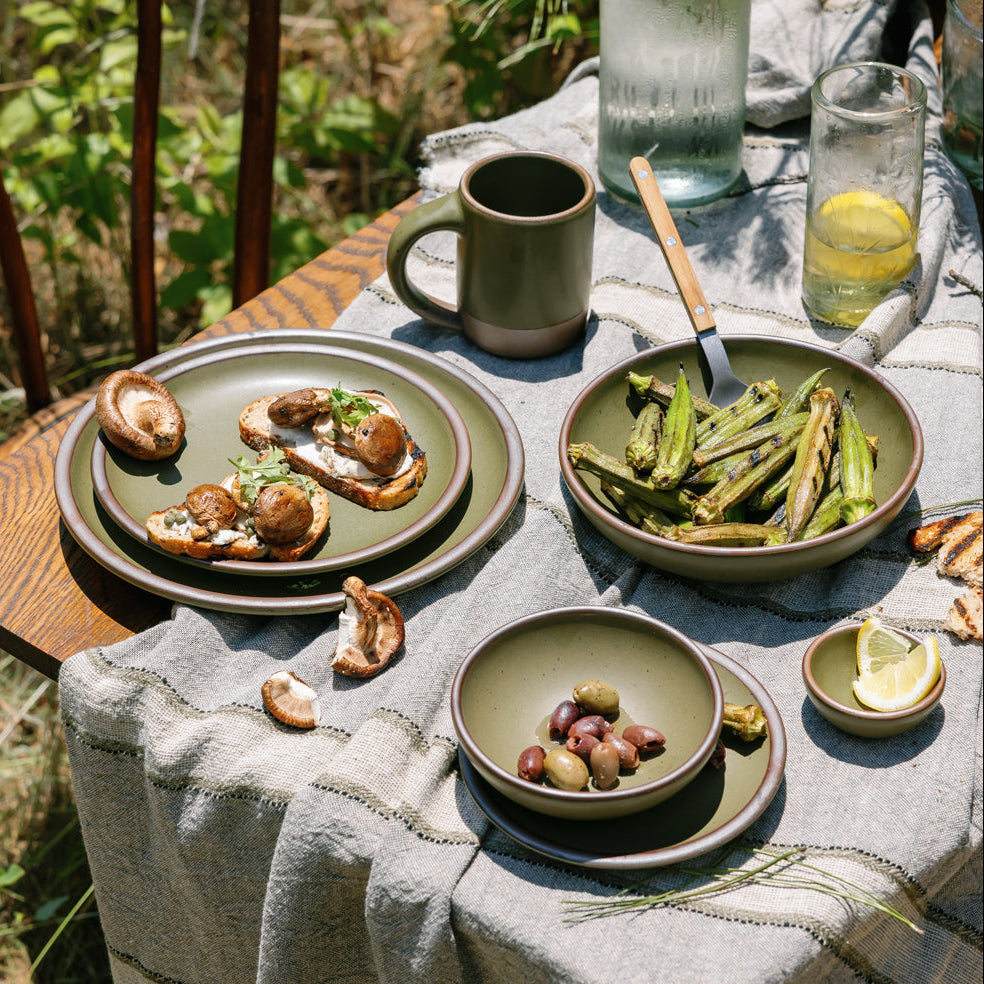Outdoor setting with a table set for a meal, featuring ceramic plates and bowls of various sizes in an olive green with food and drinks.