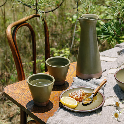 Ceramic carafe and cups in an olive green color on a wooden table outside.