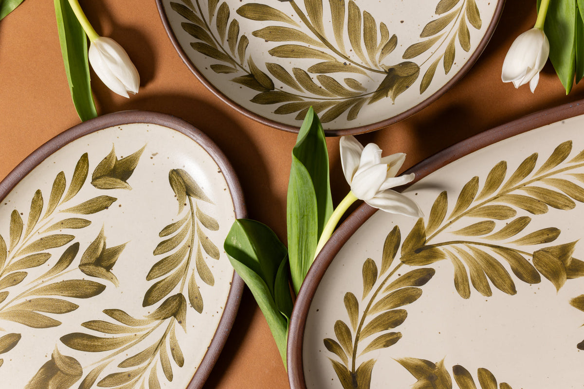 Closeup of oval ceramic platters and a shallow bowl in a warm, tan-toned, off-white color featuring iron speckles and an unglazed rim, with handpainted greenery in an olive green color, adornedwith flowers.