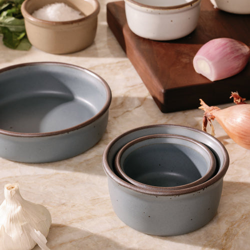 A trio of ramekins in different sizes in a blue-grey color with ungrazed rims, with a cutting board, shallot, and garlic.