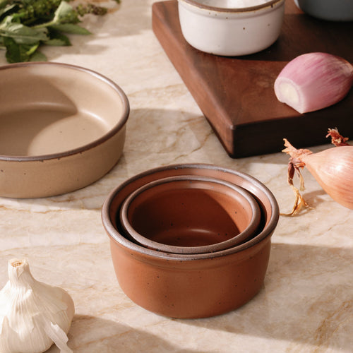 Two nesting ramekins in different sizes in a cool terracotta color with ungrazed rims, with a cutting board, shallot, and garlic.