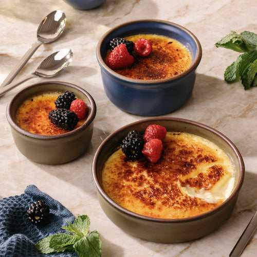 A trio of ramekins in different sizes in an olive green and blue colors with unglazed rims, filled with creme brulee and topped with berries, on a kitchen counter.