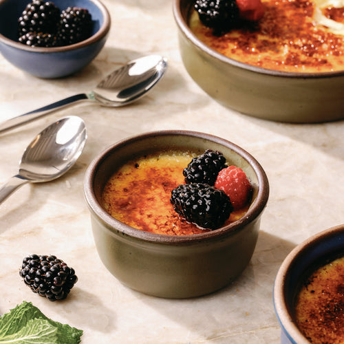 A small ramekin in an olive green color with unglazed rims, filled with creme brulee and topped with berries, on a kitchen counter.