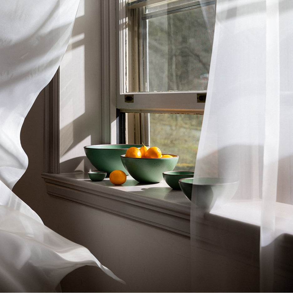 Green bowls with oranges on a windowsill with white curtains