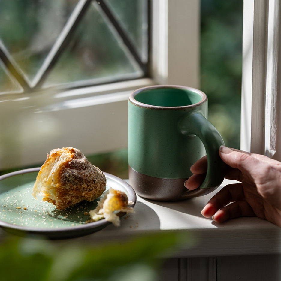 Green mug with brown base held by a hand on a windowsill with a pastry.