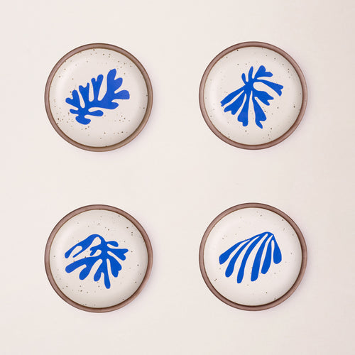 Four ceramic dessert sized plates in a warm off-white color with unglazed rim, with different frond-like cut-outs from Henri Matisse’s La Perruche et la sirène.