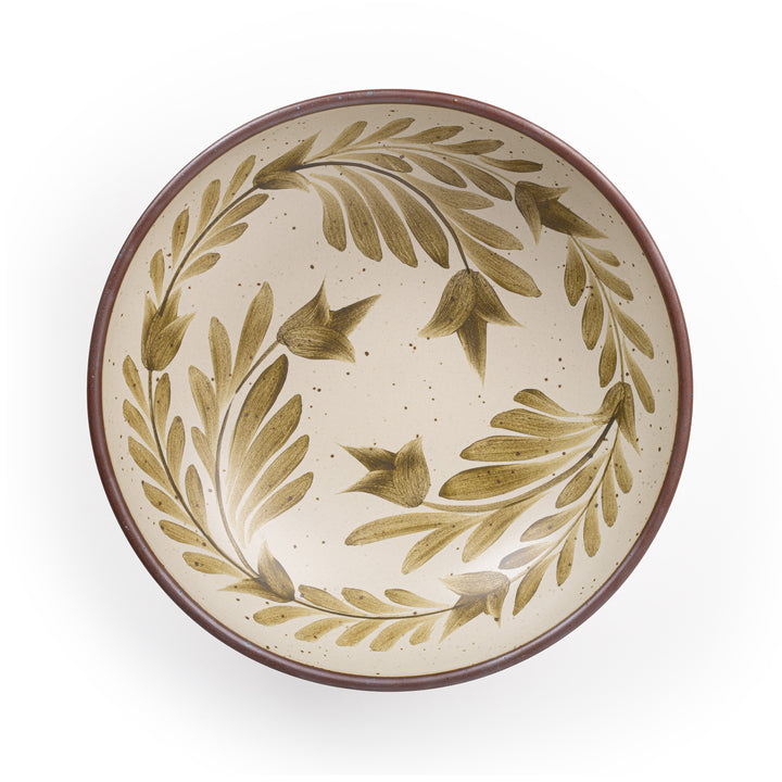 An overhead view of a large shallow serving ceramic bowl in a warm, tan-toned, off-white color featuring iron speckles and an unglazed rim, with handpainted greenery in an olive green color inside the bowl.