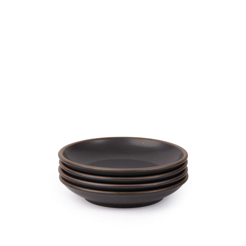 A stack of 4 small ceramic plates with a curved bowl edge in a graphite black color featuring iron speckles and an unglazed rim.