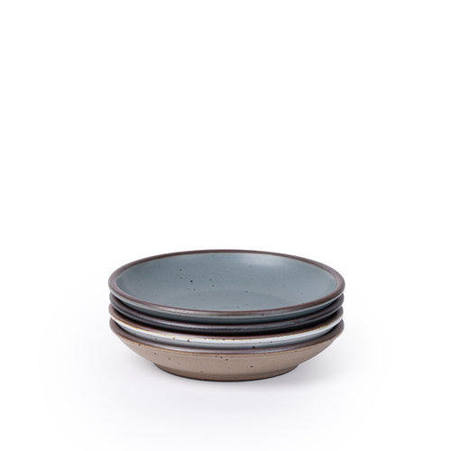 A stack of 4 small ceramic plates with a curved bowl edge featuring iron speckles and an unglazed rim in blue-grey, graphite black, cool white, and pale warm brown.