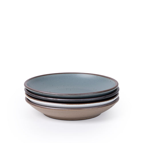 A stack of 4 medium ceramic plates with a curved bowl edge featuring iron speckles and an unglazed rim in blue-grey, graphite black, cool white, and pale warm brown.
