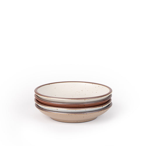 A stack of 4 small ceramic plates with a curved bowl edge featuring iron speckles and an unglazed rim in warm off-white, cool terracotta, cool white, and pale warm brown colors.