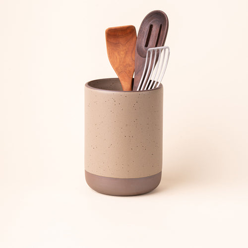 A cylindrical ceramic utensil crock and an unglazed base, in a pale warm brown color, with various kitchen utensils inside