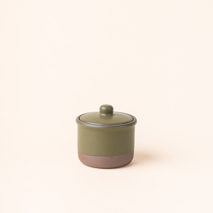 A short ceramic lidded jar in an olive green color with an unglazed base.