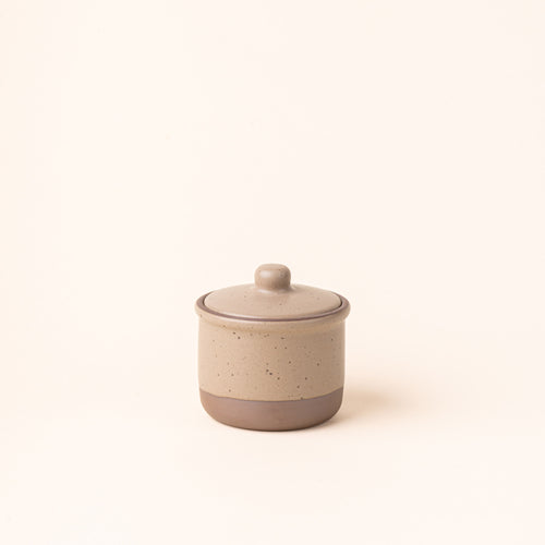A short ceramic lidded jar in a pale warm brown color with an unglazed base.