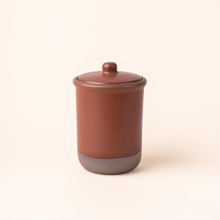 A tall ceramic lidded jar in a cool terracotta color with an unglazed base.