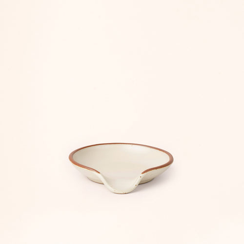A ceramic spoon rest with a curved notch in a warm off-white color with an unglazed rim.