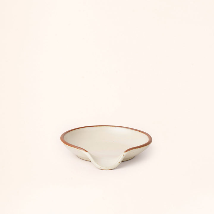 A ceramic spoon rest with a curved notch in a warm off-white color with an unglazed rim.