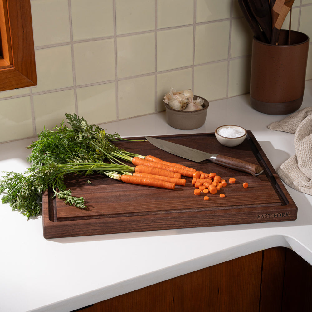 A large walnut cutting board with a juice groove, with carrots mid-cut with a knife with a knife and salt and garlic nearby.