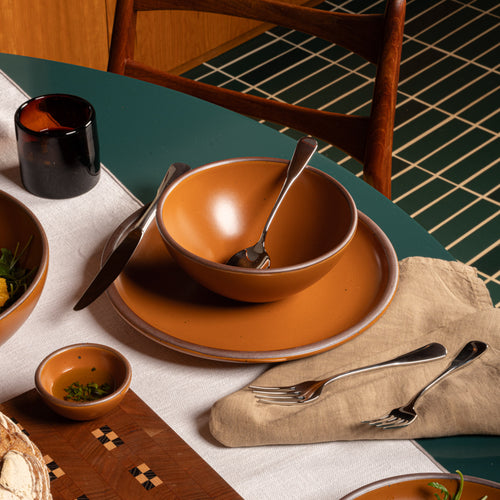A earthy orange -colored ceramic dinnerware set arranged on a dining table, including a bowl, matching plate, small side dish, and silver cutlery, styled with a beige napkin, dark candle, and wooden chair in a real interior setting.