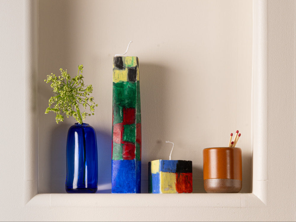A built in shelf in a wall, with hand painted rectangular tall and short candles in a colorful checkerboard pattern, a blue vase with a stem of greenery, and a small ceramic vessel with matches.