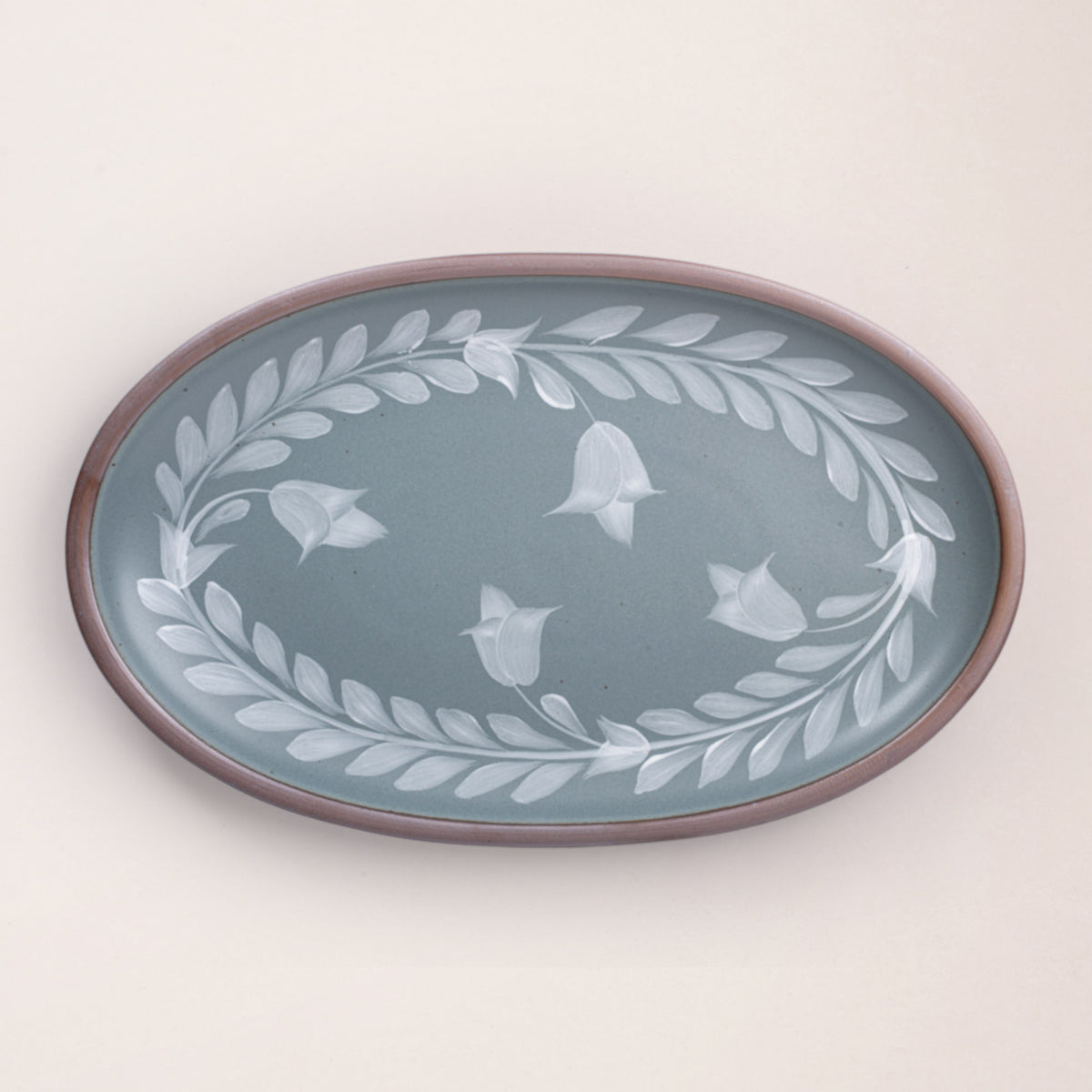 Oval ceramic platter with a hand-painted white leaf and flower border on a blue-grey background with an exposed clay rim.