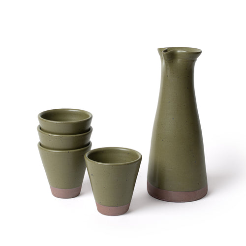 A tall ceramic carafe and stack of 4 tapered cups in an olive green color, featuring iron speckles and unglazed bases.