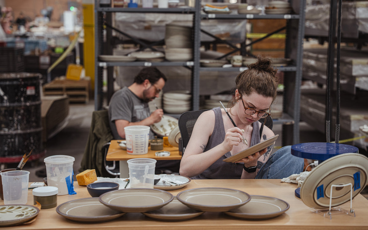 Rachel and Alex in a pottery studio sitting and painting on ceramic plates.