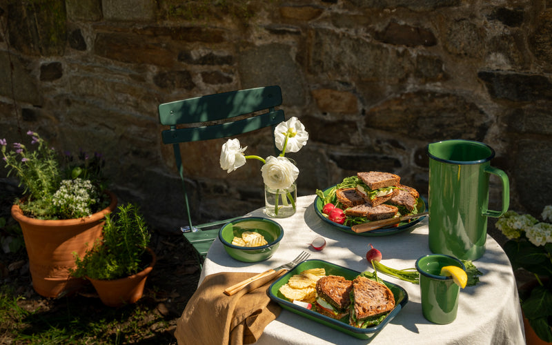 Outdoor table set with green enamelware, sandwiches, chips, radishes, and drinks, with white flowers in a vase beside a stone wall.