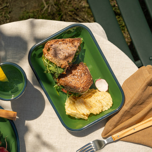 Medium rectangular baking dish in a green color with a dark green rim, filled with a sandwich and some chips on a tablecloth outside.