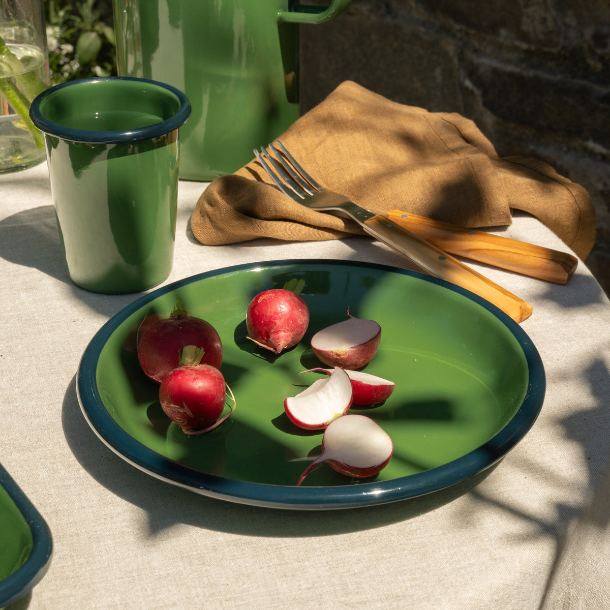 Green enamel side plate with radishes on a table with a green cup and fork.