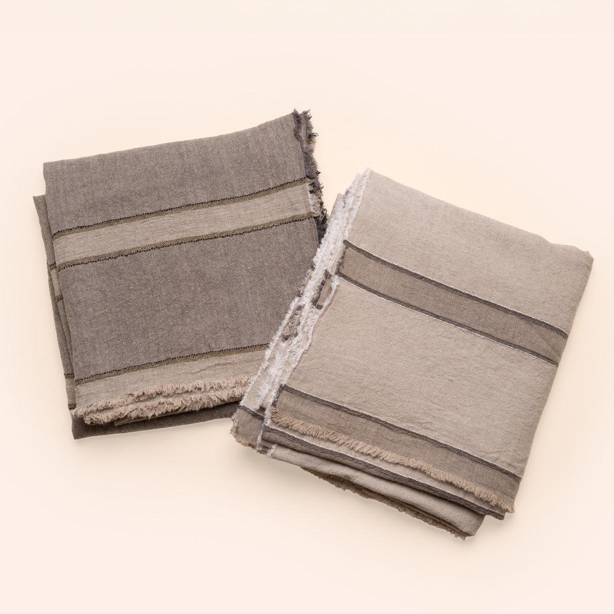 Two neutral and gray linen tablecloths with cream and olive or gray woven stripes and fringed edges, neatly folded on a clean neutral background.
