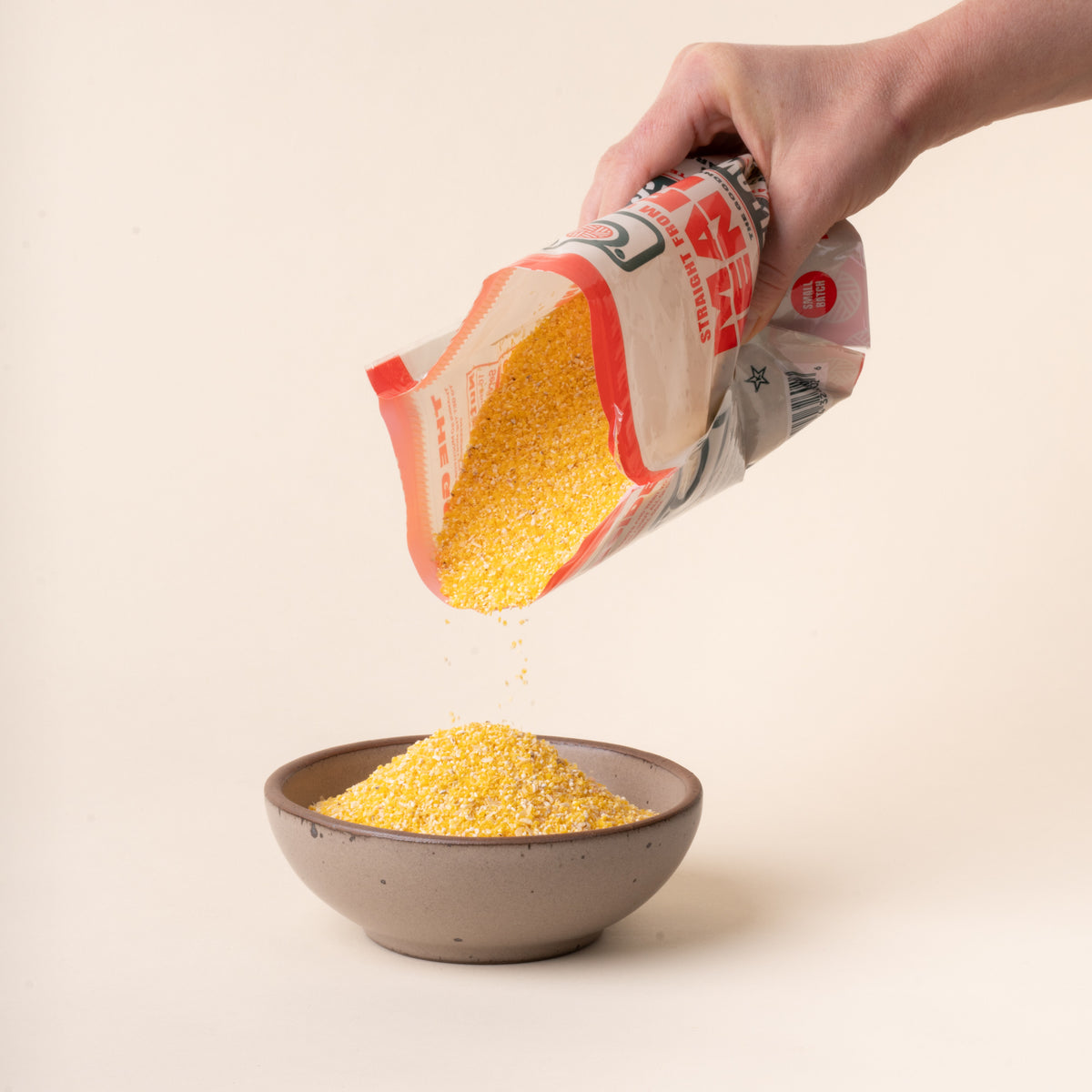 Hand pouring yellow stone-ground grits from an opened Marsh Hen Mill bag into a ceramic bowl on a plain white background.
