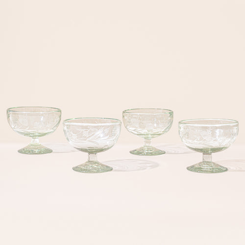 Four clear etched coupe glasses with a pedestal base and delicate botanical pattern, shown on a clean neutral background.