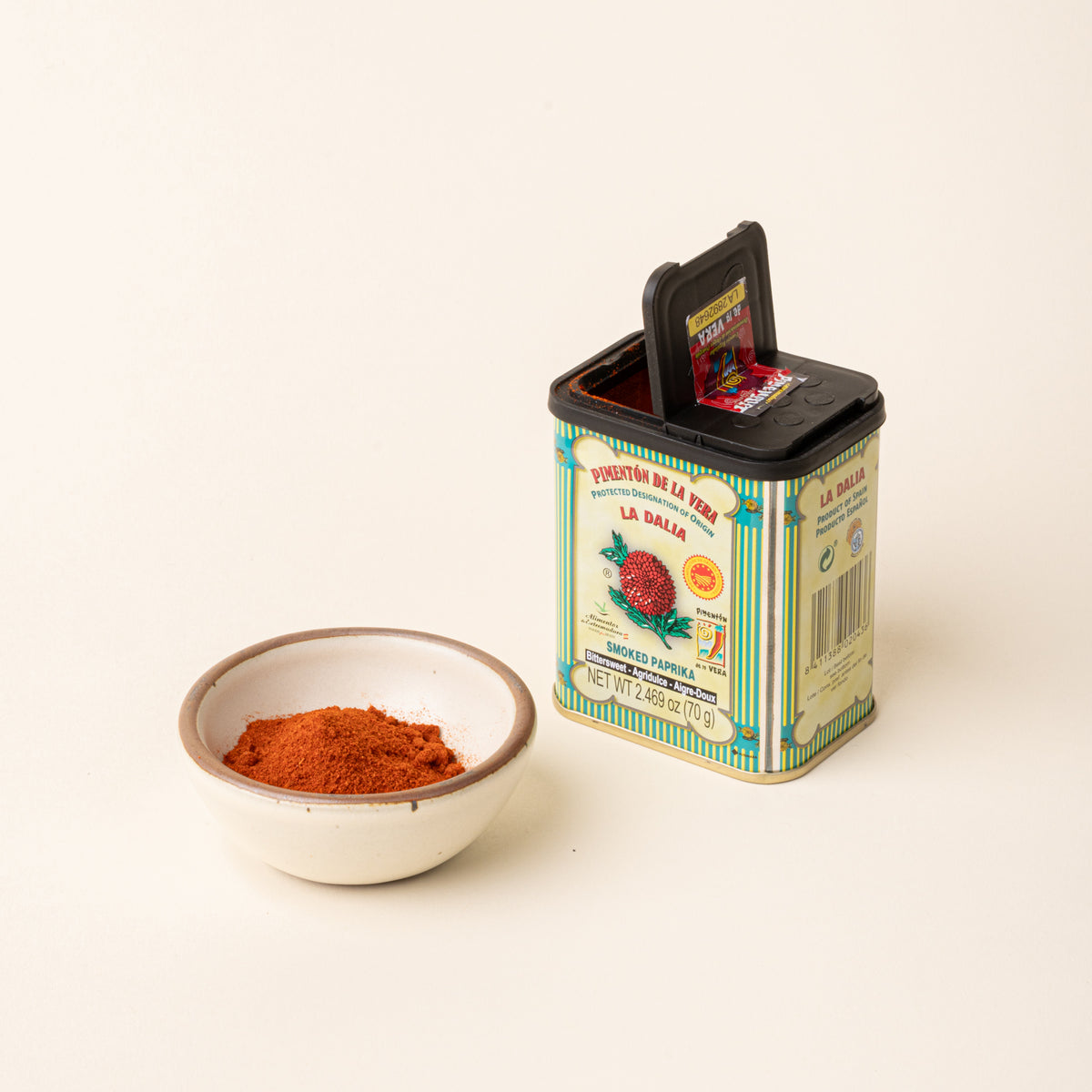 Open tin of La Dalia smoked paprika beside a small ceramic bowl filled with ground paprika on a plain white background.