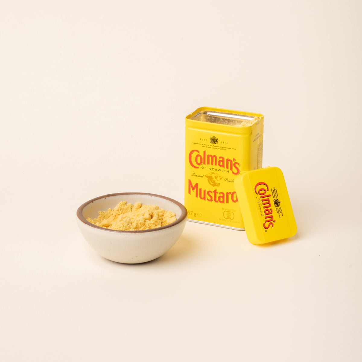 Open tin of Colman's Mustard Powder beside a ceramic bowl filled with mustard powder on a plain white background.