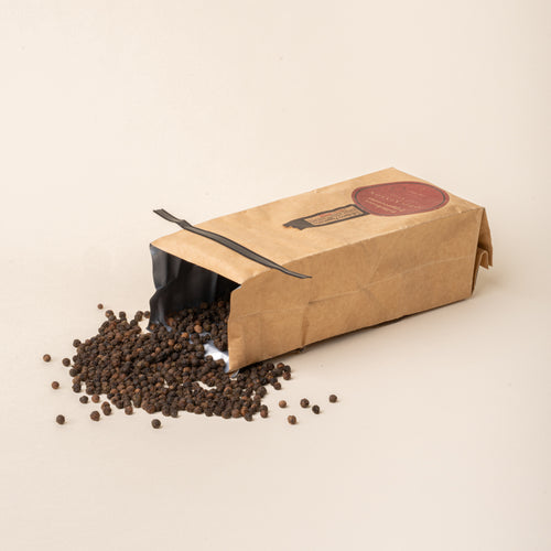 Opened bag of J.Q. Dickinson Tellicherry Peppercorns with whole black peppercorns spilled onto a plain white background.