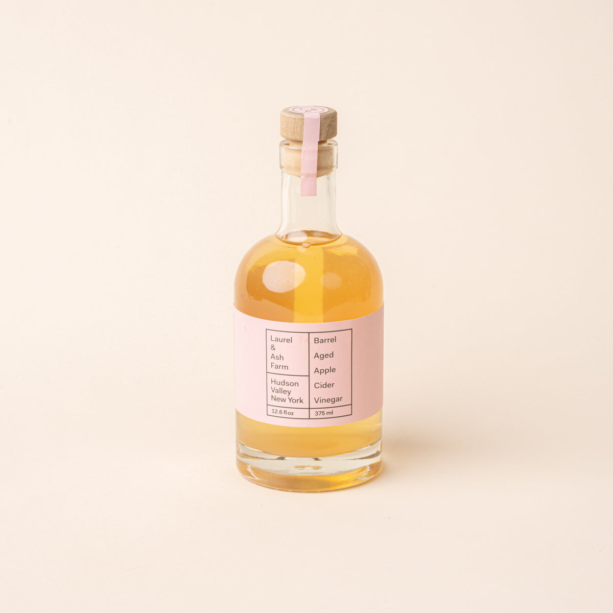 Bottle of Laurel & Ash Farm barrel-aged apple cider vinegar with a pink label and wooden stopper on a plain white background.