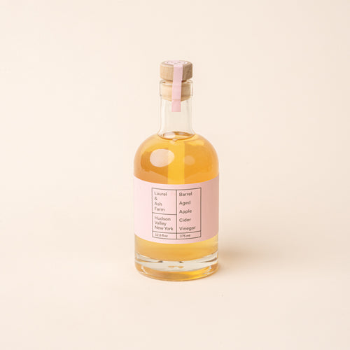 Bottle of Laurel & Ash Farm barrel-aged apple cider vinegar with a pink label and wooden stopper on a plain white background.