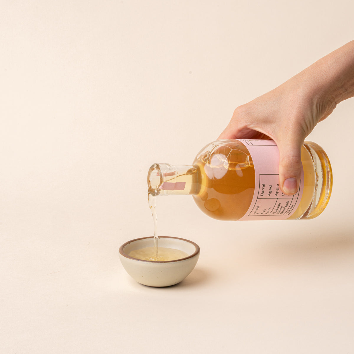 Hand pours Laurel & Ash Farm barrel-aged apple cider vinegar into a small ceramic bowl on a plain white background.