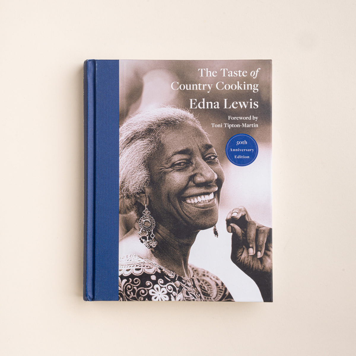 The Taste of Country Cooking book by Edna Lewis, featuring a cream-colored cover with blue decorative borders, a central portrait of the author, and red text, set against a light beige background.