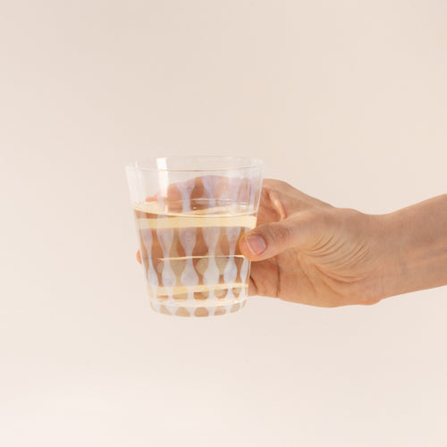 A hand hold a short clear glass with a delicate vertical wave pattern, half filled with a drink