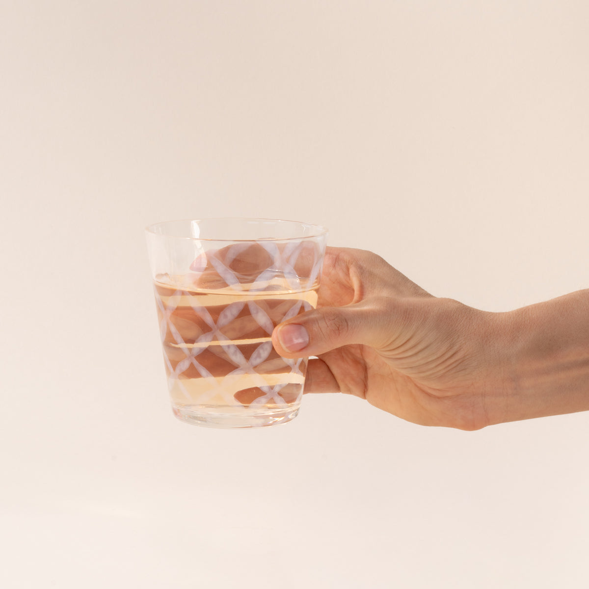 A hand holds a short clear glass with a delicate grid pattern, half filled with a drink
