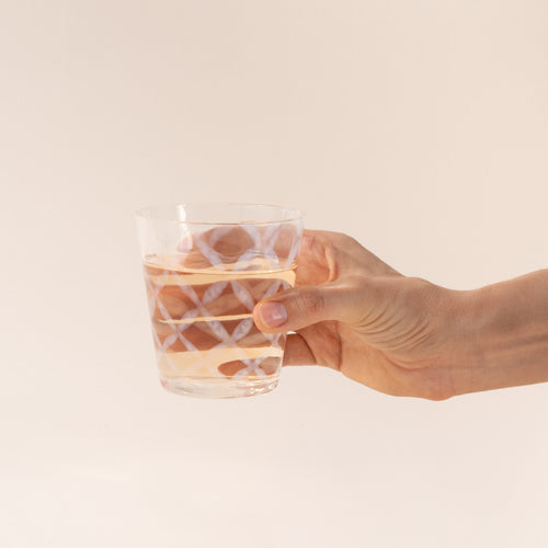 A hand holds a short clear glass with a delicate grid pattern, half filled with a drink