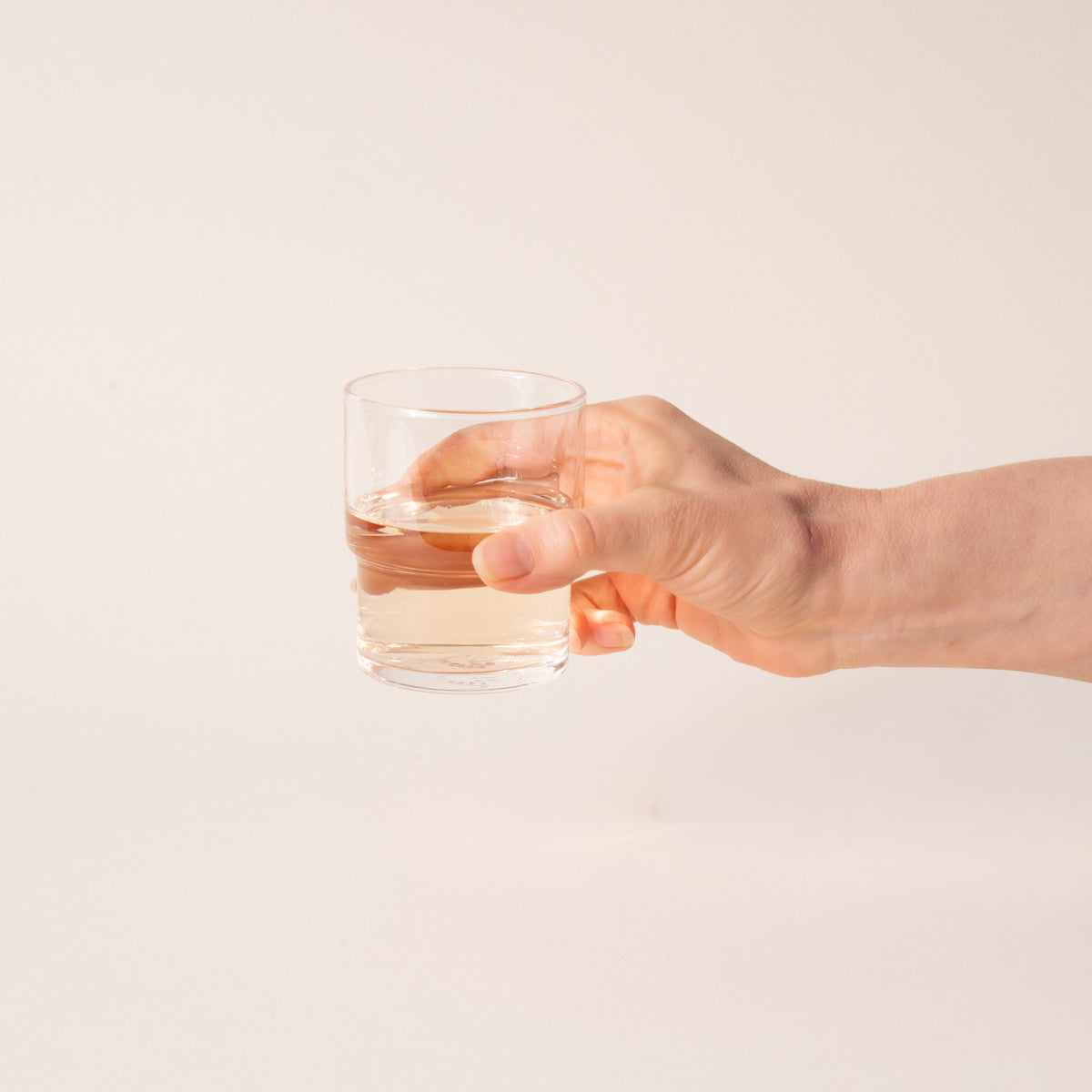 Hand holds a clear cylindrical glass with a wider top half with a narrower bottom half, filled with a drink.