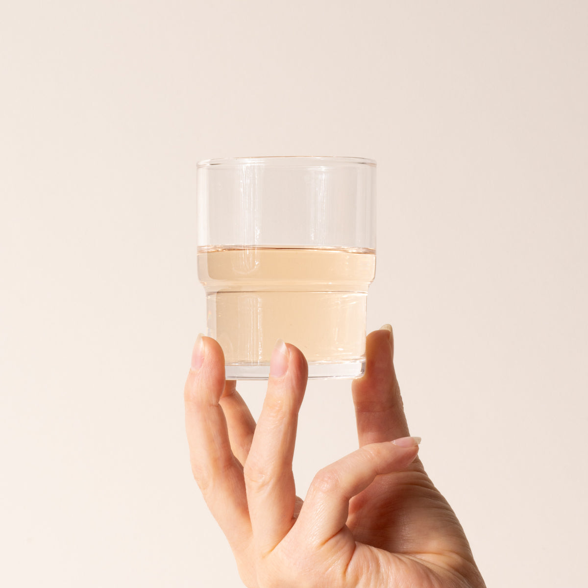 Hand holds a clear cylindrical glass with a wider top half with a narrower bottom half, filled with a drink.