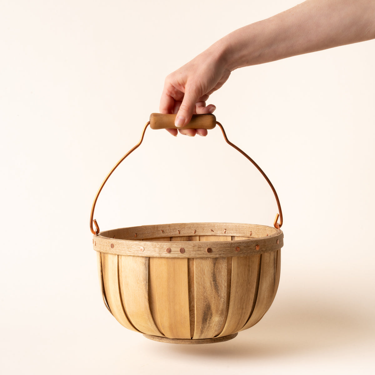 A hand holds a round wooden gathering small basket with copper handle and rivet details, shown against a light background.