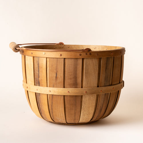 Large wooden bushel basket with copper handle, copper rivets, and alternating wood slats against a light background.