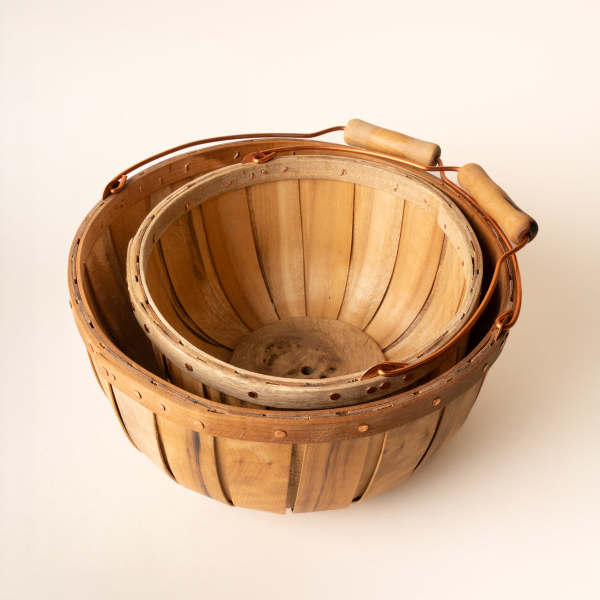 Two nested wooden bushel baskets with copper handles and rivets, showing slatted interiors and natural wood grain.