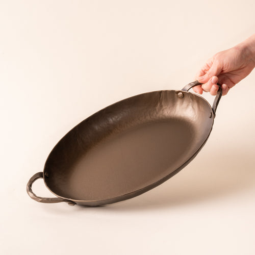 A hand tilting a hammered metal oval roasting pan with handles on both sides